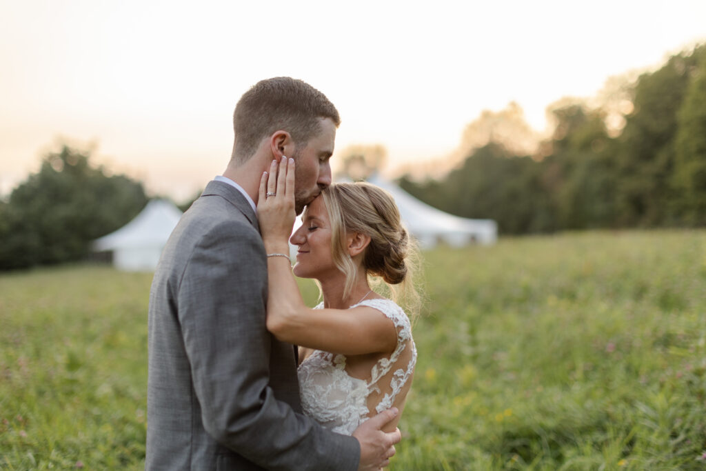 Husband kisses wife on the top of her forehead in a field at sunset. 