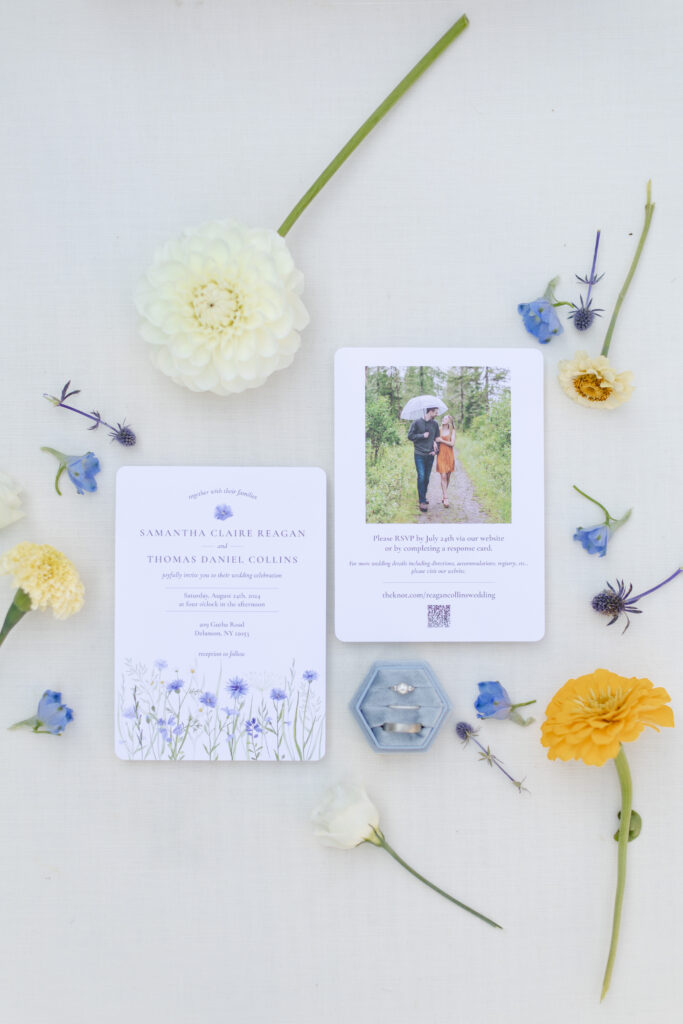 An overhead view of a flat-lay of a wedding invitation, some rings, and florals. 