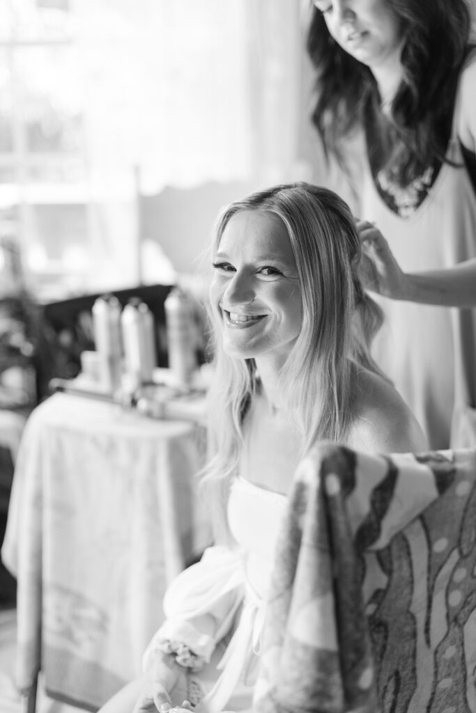 Black and white photo of the bride smiling at the camera while she gets her hair done. 