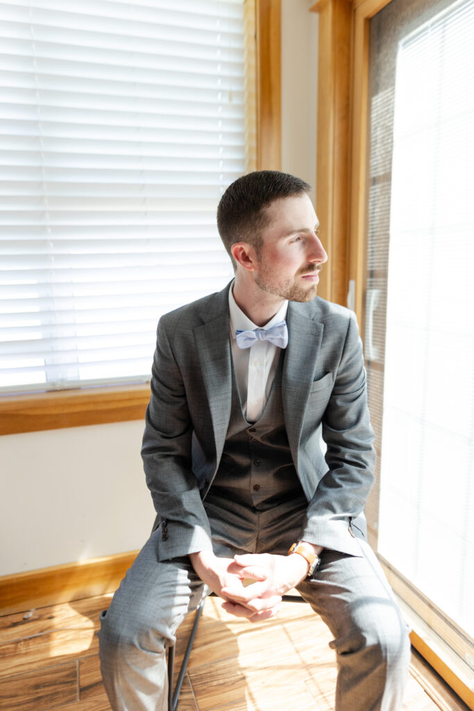 Groom sitting on a chair in his wedding suit, looking out the window. 